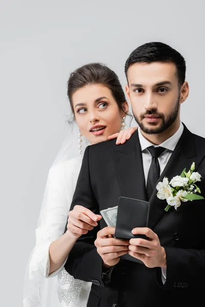 Noivo muçulmano segurando carteira com dólares perto de noiva sorridente no fundo borrado isolado no cinza — Fotografia de Stock