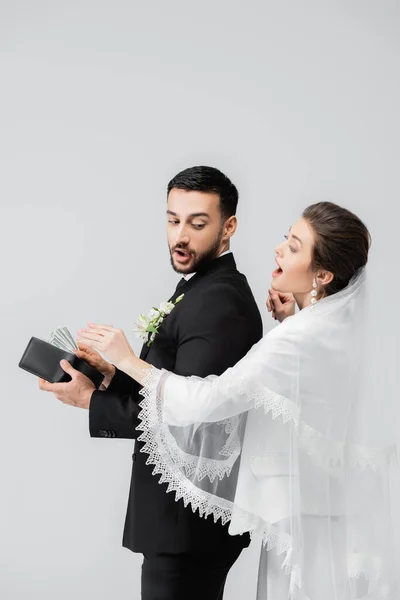 Mariée excitée debout près du marié arabe avec des dollars et portefeuille isolé sur gris — Photo de stock