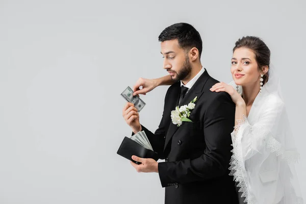 Sonriente novia tomando moneda de novio musulmán aislado en gris - foto de stock