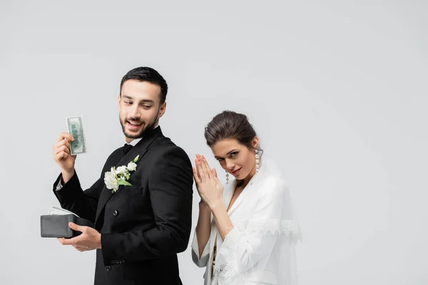 Novio musulmán sosteniendo billete de dólar cerca de novia difícil aislado en gris - foto de stock