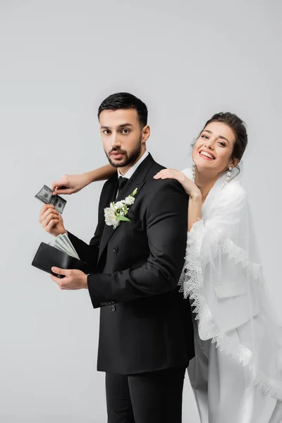 Smiling bride taking money from arabian boyfriend with wallet isolated on grey — Stock Photo