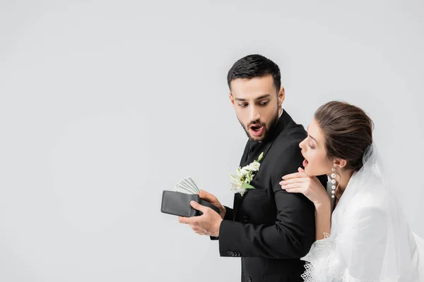 Maravilhado noivo muçulmano escondendo carteira com dinheiro perto da noiva isolado em cinza — Fotografia de Stock