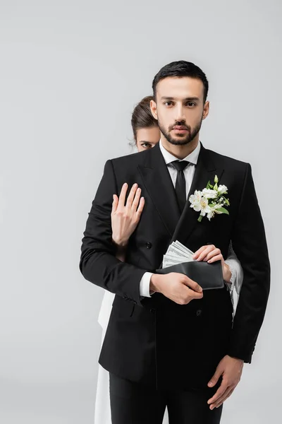 Novio musulmán mirando a la cámara mientras la novia toma el dinero de la cartera aislado en gris - foto de stock