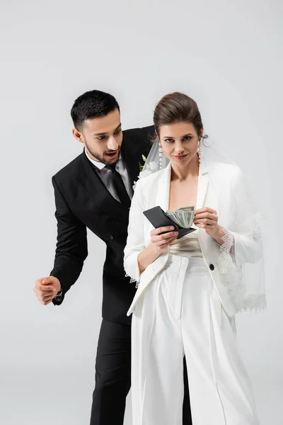 Emocionado novio árabe mirando la cartera y el dinero en efectivo en manos de la novia aislado en gris - foto de stock