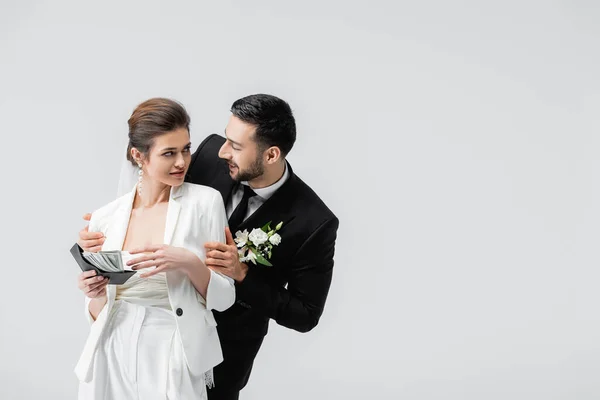 Souriant marié arabe étreignant mariée avec de l'argent et portefeuille isolé sur gris — Photo de stock