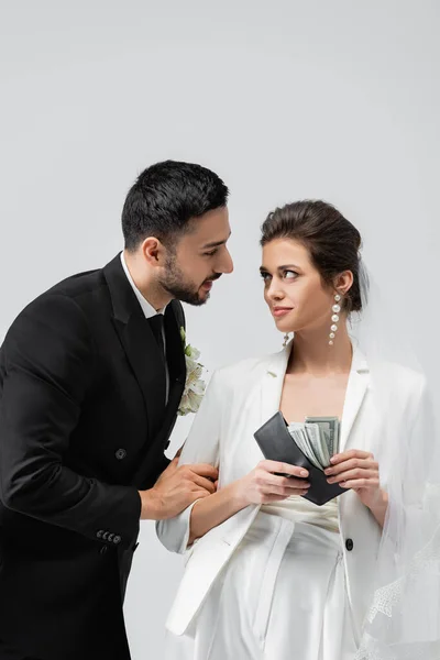 Sorrindo noiva segurando dinheiro e carteira e olhando para noivo árabe isolado em cinza — Fotografia de Stock