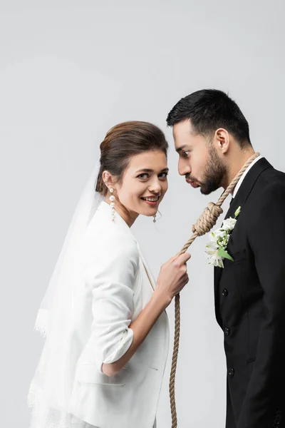 Cheerful bride holding slip knot on neck of arabian groom pouting lips isolated on grey — Stock Photo
