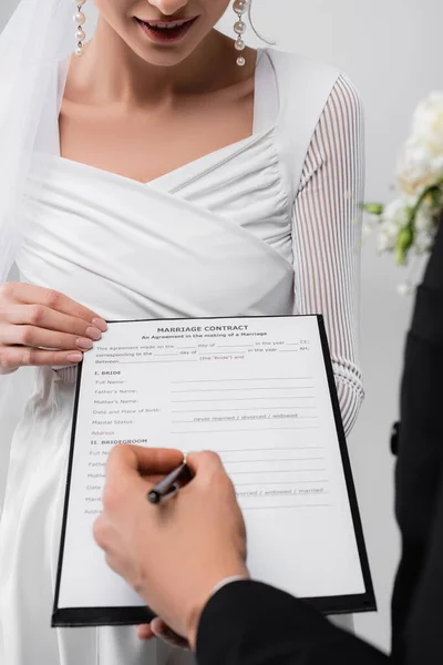 Cropped view of smiling bride holding marriage contract near groom with pen on blurred foreground isolated on grey — Stock Photo