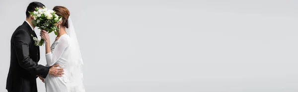 Groom and pregnant bride hiding behind wedding bouquet isolated on grey, banner — Stock Photo