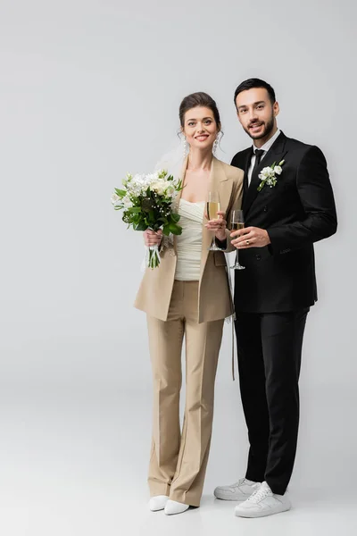 Smiling interracial newlyweds holding glasses of champagne on grey background — Stock Photo