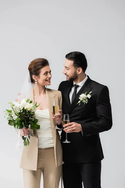 Smiling bride holding bouquet and glass of champagne near muslim groom isolated on grey — Stock Photo