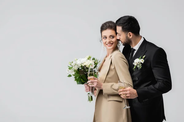 Smiling bride with wedding bouquet looking at arabian groom with champagne isolated on grey — Stock Photo