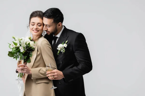 Homem árabe elegante com copo de champanhe abraçando noiva feliz segurando buquê de casamento isolado no cinza — Fotografia de Stock