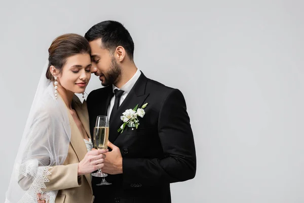 Радостная, межрасовая молодоженатая пара, стоящая с закрытыми глазами и бокалами шампанского, изолированными на сером — стоковое фото