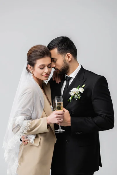 Happy, interracial newlywed couple holding champagne glasses isolated on grey — Stock Photo