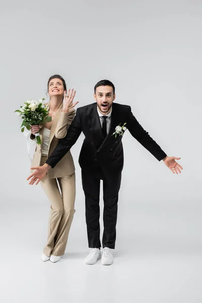 Mariée excitée montrant bague de mariage près de fiancé musulman étonné debout avec les bras ouverts sur gris — Photo de stock