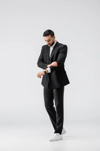 Full length view of stylish muslim man checking time on wristwatch while standing on grey — Stock Photo