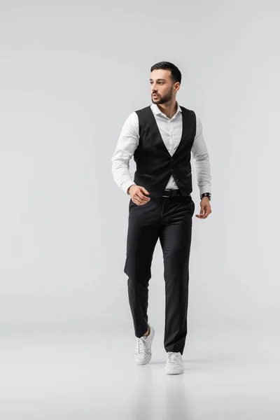Vista a tutta lunghezza dell'uomo arabo alla moda in abiti eleganti e scarpe da ginnastica che camminano sul grigio — Foto stock