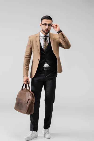 Full length view of fashionable arabian man adjusting eyeglasses while posing with leather bag on grey — Stock Photo