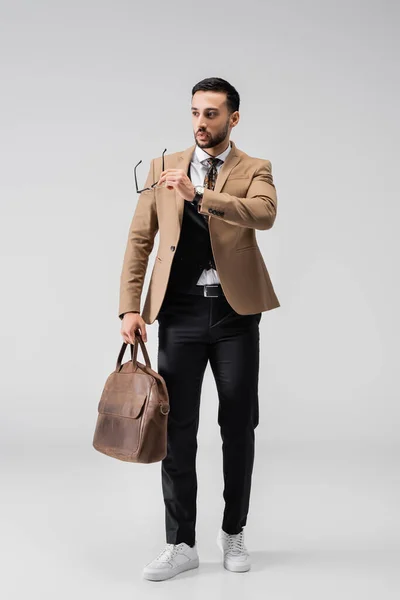 Full length view of fashionable arabian man holding eyeglasses and leather bag on grey — Stock Photo