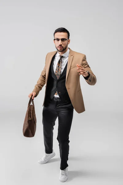 Full length view of stylish arabian man looking at camera while walking with leather bag on grey — Stock Photo