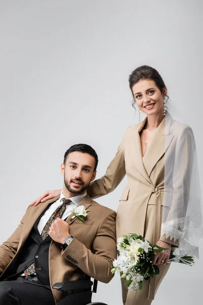 Joyful woman with wedding bouquet touching shoulder of handicapped arabian fiance isolated on grey — Stock Photo