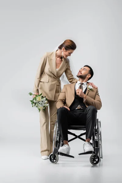 Sposa felice con bouquet da sposa sorridente a elegante, sposo musulmano handicappato sul grigio — Foto stock