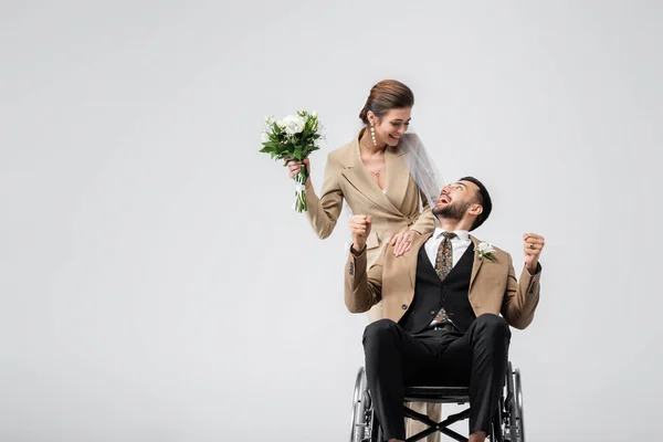 Eccitato, disabile uomo arabo mostrando vincere gesto vicino sposa elegante con bouquet di nozze isolato su grigio — Foto stock