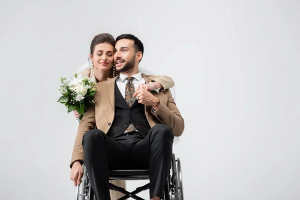 Elegant woman with wedding bouquet holding hands with handicapped arabian groom isolated on grey — Stock Photo