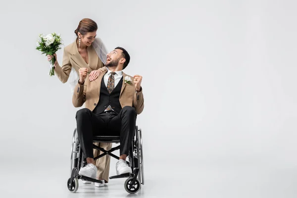Excited, handicapped arabian man showing win gesture while looking at happy bride on grey — Stock Photo