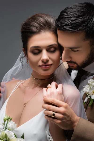 Hombre árabe tocando la mano de la novia sonriendo con los ojos cerrados aislados en gris - foto de stock