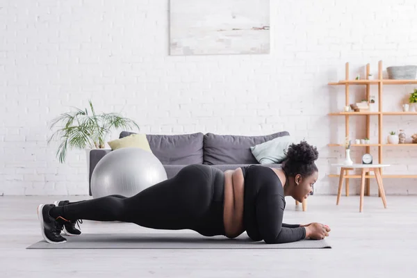 Seitenansicht einer afrikanisch-amerikanischen Plus-Size-Frau in Sportbekleidung beim Planken im Wohnzimmer — Stockfoto