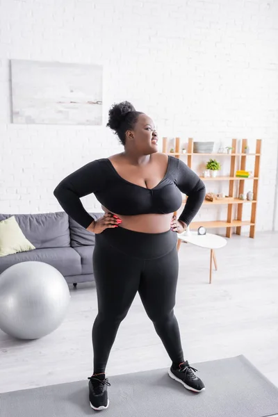 Glückliche afrikanisch-amerikanische Plus-Size-Frau in Sportbekleidung mit den Händen auf den Hüften im Wohnzimmer — Stockfoto