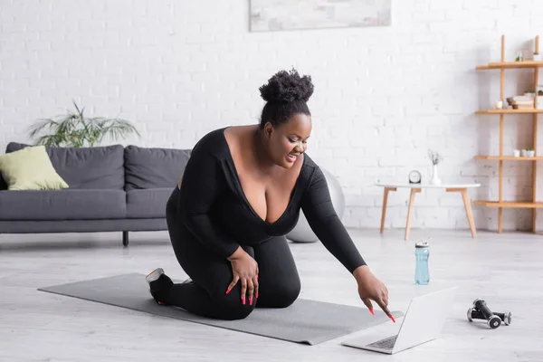 Heureux afro-américain plus taille femme regarder l'entraînement sportif sur ordinateur portable tout en étant assis sur tapis de fitness dans le salon — Photo de stock