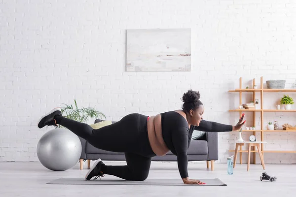 Боковой вид жизнерадостной афроамериканки plus size в спортивной одежде на фитнес-коврике — Stock Photo