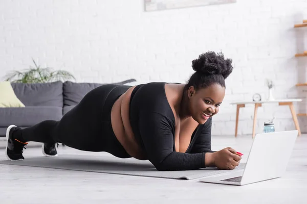 Happy african american plus size woman watching online sport training on laptop while doing plank in living room — Stock Photo