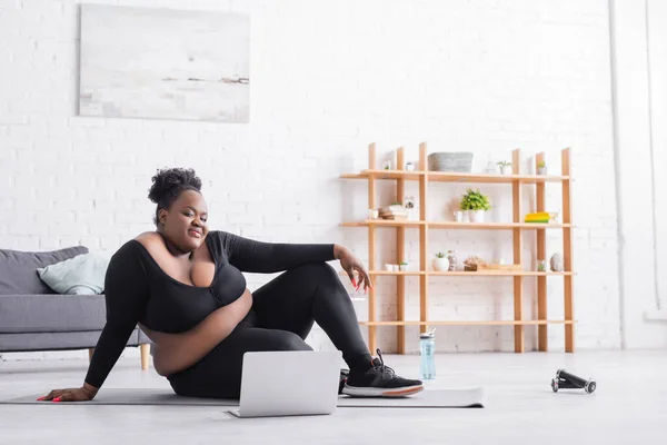 Souriant afro-américain plus taille femme regarder la formation en ligne sur ordinateur portable tout en étant assis sur tapis de fitness — Photo de stock