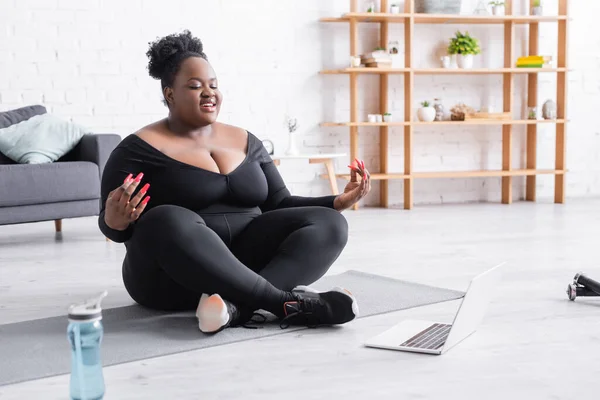 Souriant afro-américain plus taille femme regarder la formation en ligne sur ordinateur portable tout en étant assis dans la pose de yoga sur tapis de fitness — Photo de stock