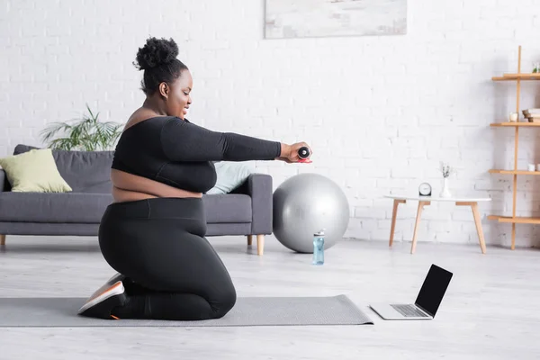 Vue latérale de la femme afro-américaine plus la taille regarder la formation en ligne sur ordinateur portable tout en exerçant avec haltère sur tapis de fitness — Photo de stock