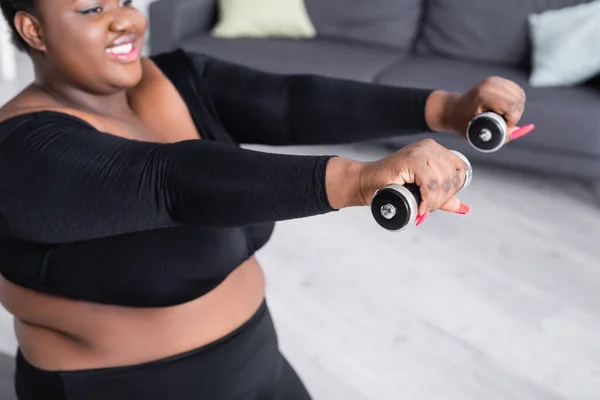 Afro-américaine floue plus la taille de la femme dans les vêtements de sport exercice avec haltères dans le salon — Photo de stock
