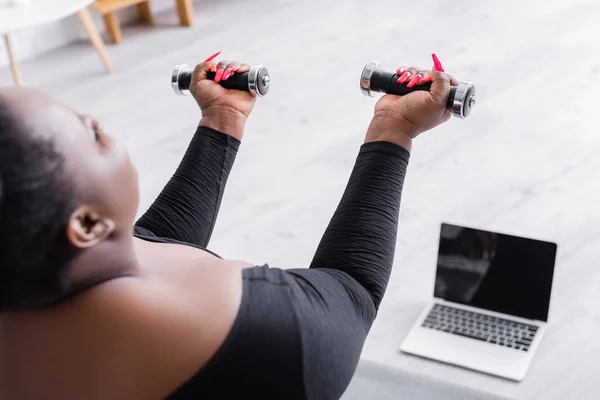 Afro-américaine floue plus la taille de la femme dans les vêtements de sport exercice avec haltères près d'un ordinateur portable dans le salon — Photo de stock