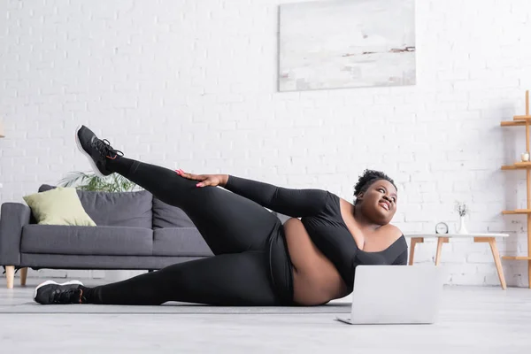 Fröhliche afrikanisch-amerikanische Plus-Size-Frau, die in der Nähe von Laptop auf Fitnessmatte trainiert — Stockfoto