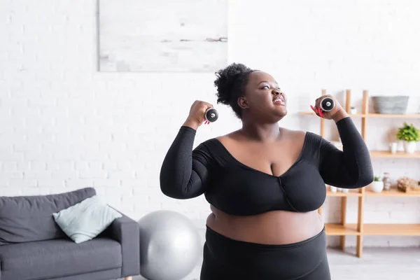 Smiling african american plus size woman in sportswear exercising with dumbbells in living room — Stock Photo