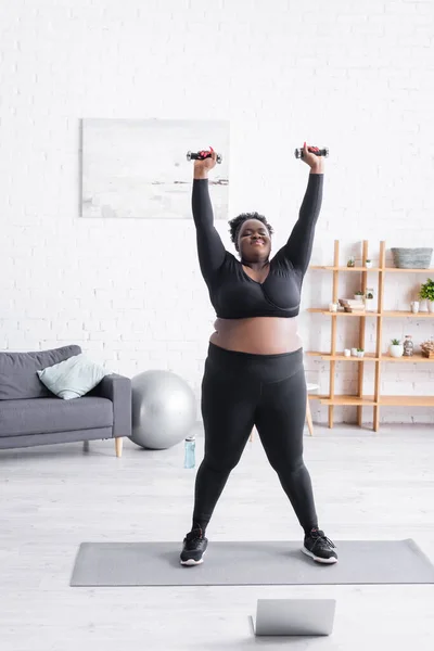 Longueur totale de femme afro-américaine plus la taille dans les vêtements de sport exercice avec haltères dans le salon — Photo de stock