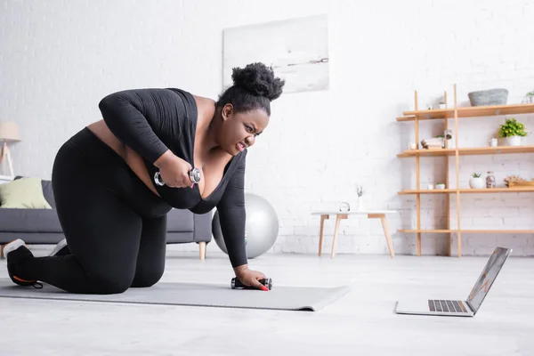 Angespannte afrikanisch-amerikanische Plus-Size-Frau in Sportbekleidung, die mit Hanteln trainiert und auf Laptop schaut — Stockfoto