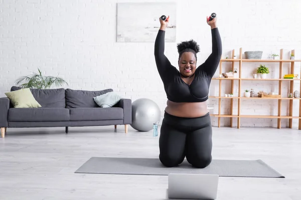 Heureux afro-américain surdimensionné femme en vêtements de sport exercice avec haltères près d'un ordinateur portable — Photo de stock