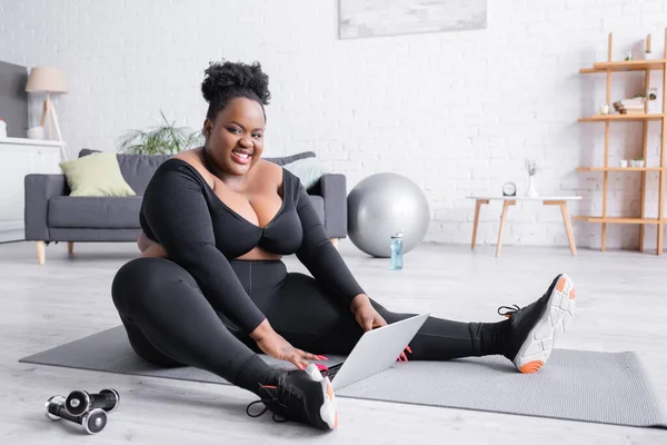 Glückliche afrikanisch-amerikanische Plus-Size-Frau in Sportbekleidung sitzt auf Fitnessmatte mit Laptop — Stockfoto