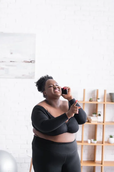 Glückliche afrikanisch-amerikanische Plus-Size-Frau in Sportbekleidung, die auf dem Smartphone spricht und mit dem Finger zeigt — Stockfoto