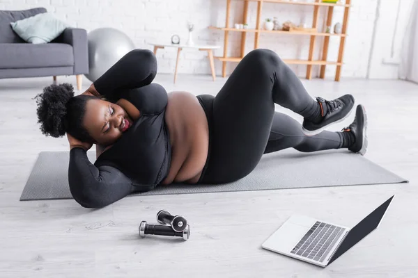 Heureux afro-américain plus taille femme en vêtements de sport travailler sur tapis de remise en forme et en regardant ordinateur portable — Photo de stock