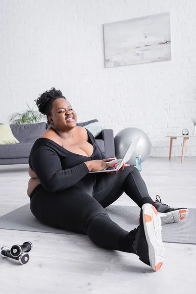 Heureux afro-américain plus taille femme en vêtements de sport tenant ordinateur portable et assis sur tapis de fitness — Photo de stock